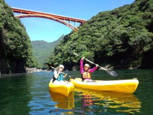 緑の木に覆われた山の間にかかる赤いアーチ橋を背景に、穏やかな川の上で、黄色いカヤックに乗った二人の女性がパドルを高く掲げながら笑顔でレジャーを楽しんでいる写真