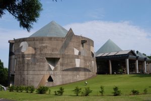 手入れされた芝生の敷地に小さな木が一列に植えられ、その背後に円筒形の打ちっ放しコンクリートの壁に幾何学模様が施され、緑色の円錐形の屋根を持つ大きな建物が建っている写真