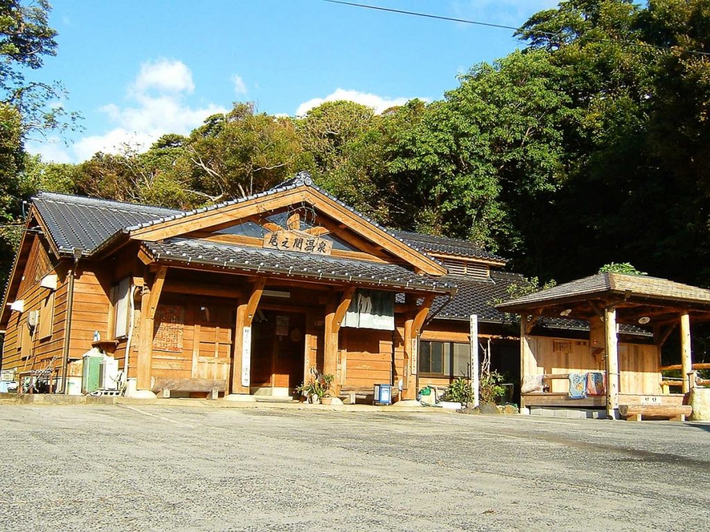 緑の森の前に尾之間温泉と書かれた看板を掲げた瓦屋根の重厚な木造建築が建ち、右手前には小さな東屋があり、手前には砂利敷きの広場がある尾之間温泉の外観写真