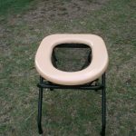 芝生の上に黒い金属製の折りたたみ脚を持ち、中央に穴の開いたベージュ色の便座が取り付けられた簡易携帯トイレ椅子の写真
