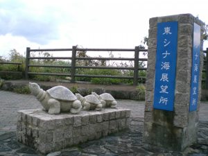 「東シナ海展望所」の青い標識が設置された刻まれた石柱の横に、四角い石の台座の上に三匹の石でできた亀の像が左を向いて並んでおり、背景には木製の柵と野草が見える展望所の写真