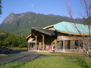 切り立った岩肌の崖が特徴的な緑の山と森を背景に、丸みを帯びた黄緑色の屋根のぽん・たん館の建物が建っている写真