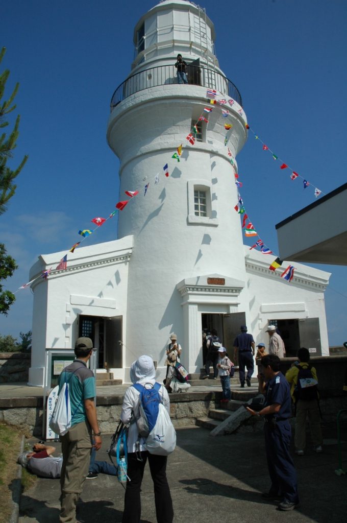 青空の下で複数の人々が出入りや見学をし、周囲にカラフルな旗が飾られている白い屋久島灯台の写真