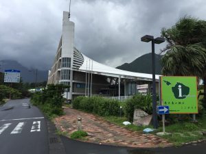 屋久島の島のシルエットの中央に「i」の文字が書かれた緑色の案内板が歩道沿いに立っており、その奥には、特徴的な白い帆のような屋根と壁を持つ近代的な建物が建っている写真
