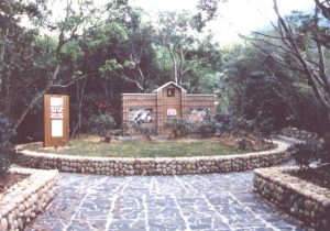 木々に囲まれた石畳の庭に、石垣の丸い花壇があり、中には芝生と低木の植栽、レンガつくりの小さな建物、左側に案内板のような看板が設置されている写真