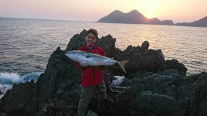 岩場の海岸で釣り竿を背後に置き、夕日を背景に海を望む中、大きな魚を両手で持って立っている人物が写り、奥には島影が見える釣果記念の写真