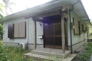 薄い灰色の壁の平屋で、玄関には格子状の引き戸がありその前には木造の柱で支えられた庇、窓には茶色の雨戸が付いた民家の外観写真