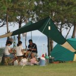 海が見えるキャンプ場で、テントとタープテントを張り、椅子に座ってキャンプを楽しむ3人の人物の写真