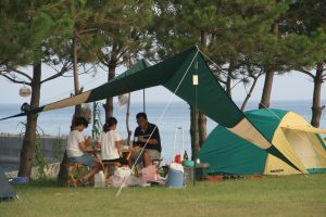 海が見えるキャンプ場で、テントとタープテントを張り、椅子に座ってキャンプを楽しむ3人の人物の写真