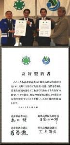 屋内の会場で、スーツやジャケットを着用した4人の男性が整列し、それぞれが白い証書を両手で持ち、両サイドの壁には自治体の旗のようなものが掲げられている写真と、その下に青森県青森市と鹿児島県屋久島町との友好盟約書の本文と関係者の署名が記された書面が合成された、盟約締結の様子を伝える写真