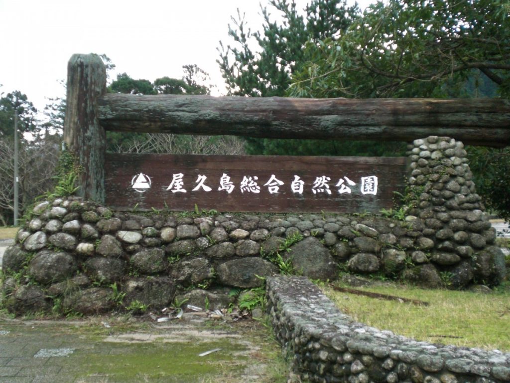 低い石垣の上に、横木と柱が載った木製の看板が設置されており、白文字で「屋久島総合自然公園」と書かれている、公園入り口の写真
