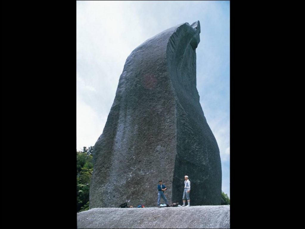 大きく切り立って垂直にそびえる濃い灰色の巨大な天柱石の下に2人の人物が立っている写真