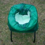 芝生の上に黒い金属製の脚を持つ簡易トイレ椅子が置かれ、便座部分には緑色の防水袋が装着され内部に白い吸水シートが見える携帯トイレの写真