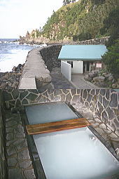 岩場の海岸に面して作られた、石垣造りの枠組みを持つ長方形の露天風呂と、その奥にある休憩所のような小さな建物を上から見下ろすように撮影した写真