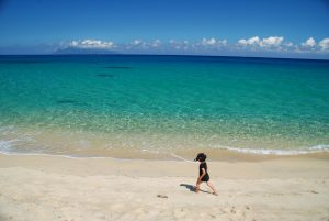 水平線に遠くの島影が見える青い海と白い砂浜の波打ち際を、黒い水着を着てゴーグルをつけた子供が一人で歩いている夏のビーチの風景写真