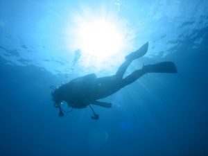 海面から太陽の光が降り注ぎ、1人のダイバーがスキューバダイビングをしているシルエットが見える海中写真