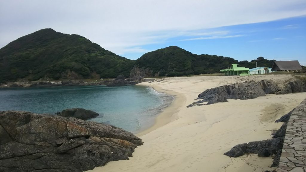 ところどころに大きな岩がある白い砂浜の前に透き通った海、奥には緑の木々に覆われた山並みが見える写真