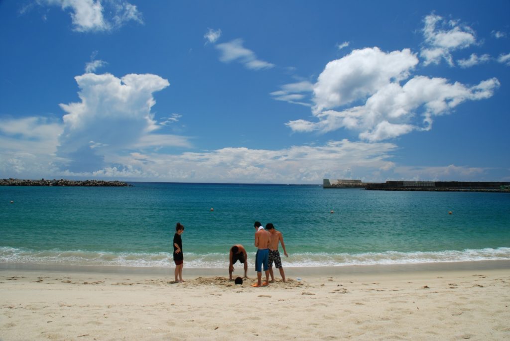 青空に大きな入道雲が広がり、防波堤が見える穏やかな海の手前にある白い砂浜で、水着姿の4人の若者が輪になって足元の砂を見ている写真