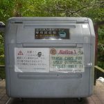 木々に囲まれた屋外に設置された大型の灰色プラスチック製廃棄ボックスで、正面には「使用済み携帯トイレ専用回収箱」など日本語と英語で注意書きが印字された白い案内板が貼られた専用ゴミ箱の写真