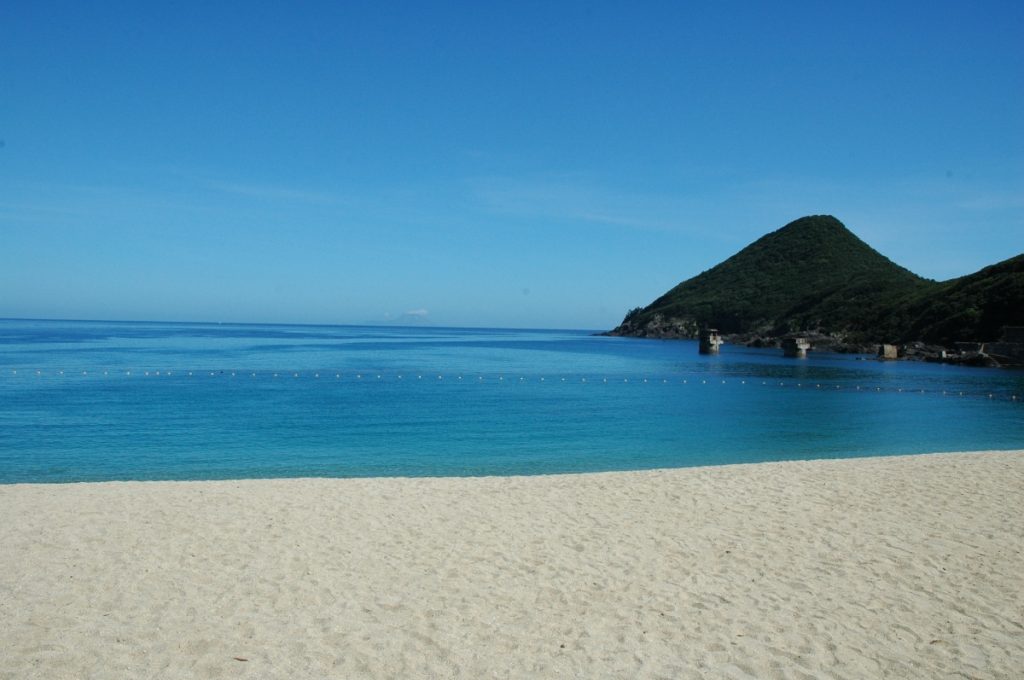 雲ひとつない鮮やかな青空の下、手前に広がる白い砂浜とエメラルドグリーンから深い青へ変化する海、その海上にはブイが列をなし右奥には緑の山が海へ突き出している海岸の風景写真