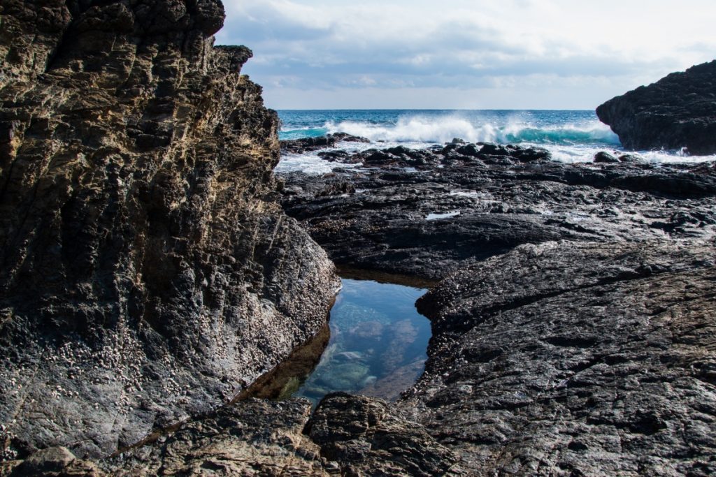 岩場に囲まれた潮だまりが中央にあり、奥には白波が立つ青い海と雲が広がる空が見える写真