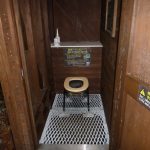 木製の壁に囲まれた屋内に白い格子状の床が敷かれ、中央に黒い脚に丸い便座を乗せた簡易トイレが設置され、奥の壁には説明書きとトイレットペーパーが置かれたトイレの写真