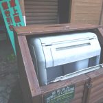 木製の囲いの中に灰色の大型廃棄ボックスが収められ、前面には「使用済み携帯トイレ回収箱」などの注意書きが貼られた、屋外設置型ゴミ箱の写真