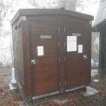 霧が立ち込める屋外に設置された濃い茶色の木製二連式トイレ小屋で、中央の扉に白い案内板が貼られ、両扉に白地に黒で「空き」と書かれた札が付いた公衆トイレの写真