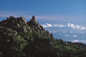 澄み渡る青空の下、緑の低木に覆われた山肌に巨大な岩塊がいくつも突き出し、遠くの空に浮かぶ白い雲と共に高山の開放的で荒々しい自然を描き出している風景写真