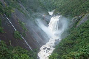 左側に広がる巨大で滑らかな一枚岩の斜面と深い緑の木々に挟まれたV字谷の奥から、白い水煙を上げながら激しく流れ落ちる滝が、圧倒的な自然のスケールを感じさせる風景写真