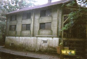 木々に囲まれた敷地に建つ二階建ての古い木造で、窓が並ぶシンプルな構造の山小屋の写真