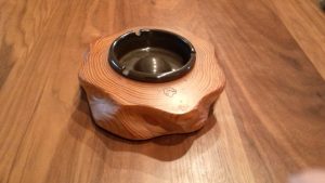 木目の美しいテーブルの上に置かれた、明るい茶色の天然木を削り出して作られた波打つ形状が特徴的な灰皿で、中央には黒い受け皿がはめ込まれ、表面の年輪模様が木の温もりを感じさせる木工製品の写真