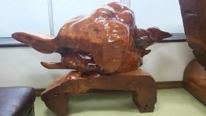 室内の台座の上に飾られた、艶やかに磨き上げられた茶色の木材が持つ複雑な凹凸や曲線の自然な造形美を活かした、独特な形状の迫力ある大きな木工芸品の写真