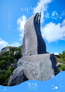 空に向かって巨大な一枚岩がそそり立つ風景を、下部から見上げるように捉え、その岩の麓に立つ人物の姿が対比された、「今、私が旅する理由」という文字入った屋久島の観光ポスター