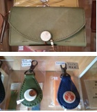 （上）薄いカーキ色で中央に丸い金具がついた革製長財布の写真、（下）丸いチャームが中央に取り付けられ、片方の端に金具がついた、丸みを帯びた形の、緑と青の小さな革製のキーホルダーが2つ並んだ写真