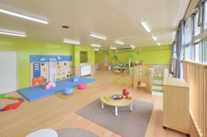 黄緑色の壁で右側に窓がある室内に、プレイマットが敷かれた遊び場や小さな遊具、テーブルなどが置かれているプレイルームの写真
