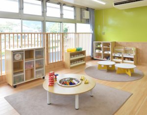 大きな窓の前には木製の安全柵が設置され、窓や壁の前にはおもちゃが収納された棚などが並び、カーペットの上に大小3つのテーブルが並べられたプレイルームの写真