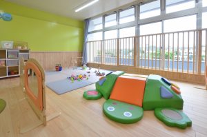 安全柵が設置された大きな窓と緑色の壁、木目調の床の室内に、カメの形をした大きなクッション遊具やおもちゃが置かれたプレイルームの写真