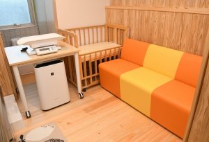 木目調の壁と床に囲まれた一室の角に、オレンジと黄色の二人掛けソファ、体重計が置かれた作業台、木製ベビーベッドと空気清浄機が設置されている授乳室の写真