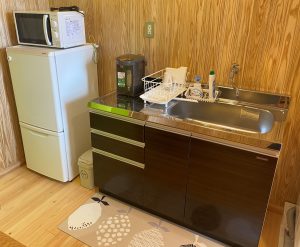 木目調の壁と床の室内の隅に、小さな冷蔵庫の上に電子レンジが置かれ、横にシンク付きの濃い茶色のシステムキッチンが設置されている写真