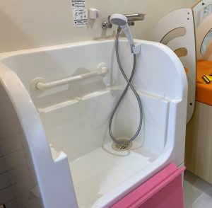 片側に手すりが付いた、底に排水口があり上にシャワーヘッドが付いている、ピンク色のドアが付いた白いシャワーシンクの写真