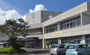 グレーの外壁の2階建てで中央に高い塔のある建物の入り口から左側の棟に、「ぽかぽかの木」の看板が設置され、手前に2台の車が停まっている写真