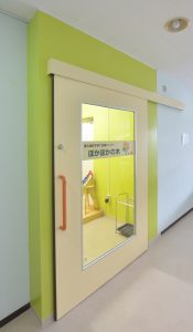 中央のガラス部分に「ぽかぽかの木」と表示が貼られた黄緑色の壁に設置された薄い黄色のドアの写真