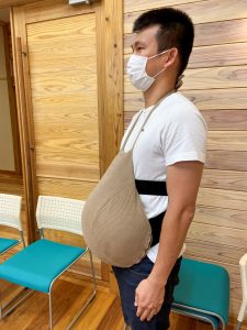 妊婦のおなかのような膨らみのベージュ色の袋を、首と腰で支えるように固定して装着した男性を横から写した写真